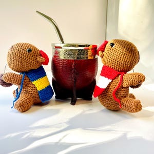 Op de afbeelding: Twee bruine, gehaakte poppen met kleurrijke sjaals zitten naast een rode yerba mate kalebas met een zilveren rand en rietje. De poppen hebben rode oren. De kalebas is ongeveer 10 cm hoog.