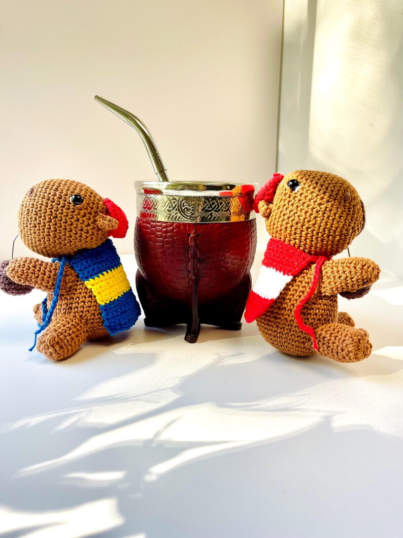 Op de afbeelding: Twee bruine, gehaakte poppen met kleurrijke sjaals zitten naast een rode yerba mate kalebas met een zilveren rand en rietje. De poppen hebben rode oren. De kalebas is ongeveer 10 cm hoog.