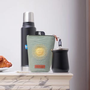 Op de afbeelding: Een olijfgroene ge&iuml;soleerde bekerhouder met een gouden zonontwerp, een zwarte thermoskan en een zwarte yerba mate-beker met een metalen rietje. Een croissant ligt op een wit bord op de achtergrond.