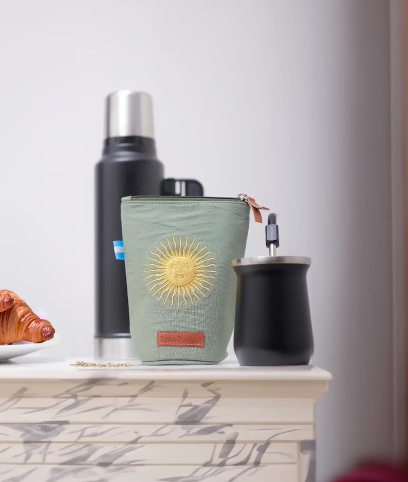 Op de afbeelding: Een olijfgroene ge&iuml;soleerde bekerhouder met een gouden zonontwerp, een zwarte thermoskan en een zwarte yerba mate-beker met een metalen rietje. Een croissant ligt op een wit bord op de achtergrond.