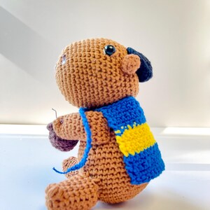 Op de afbeelding: Een gehaakte bruine berenpop met een blauw-gele sjaal. De beer heeft zwarte ogen en een kleine, donkerblauwe hoed. De handgemaakte pop heeft een grillig, vriendelijk uiterlijk.