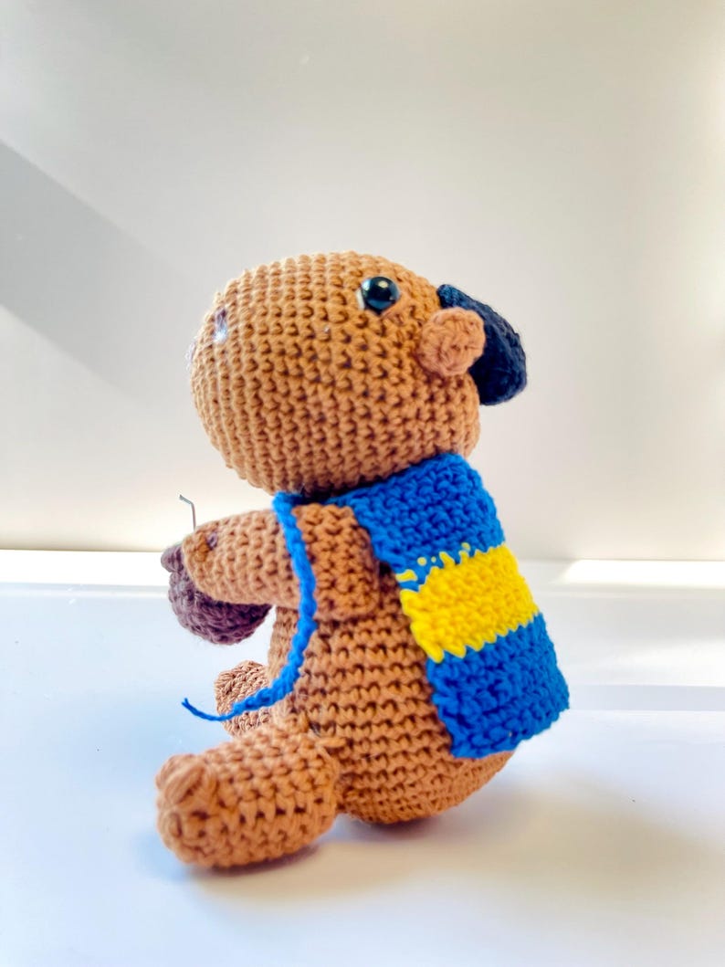 Op de afbeelding: Een gehaakte bruine berenpop met een blauw-gele sjaal. De beer heeft zwarte ogen en een kleine, donkerblauwe hoed. De handgemaakte pop heeft een grillig, vriendelijk uiterlijk.