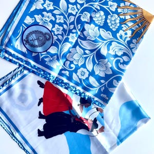 May include: Two square silk scarves. One features a blue and white floral pattern with a circular emblem and a sun design. The other scarf has a white background with a blue wave design and a graphic of two people dancing in red and black.