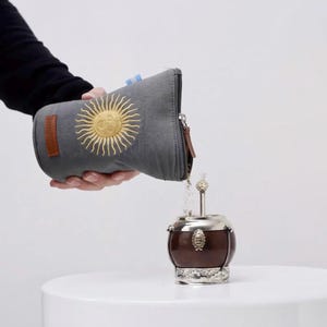 Op de afbeelding: Een grijze etui met een gouden zonnembleem giet inhoud in een bruine en zilveren yerba mate kalebas. De kalebas staat op een wit oppervlak. De etui heeft een bruin leren patch en een rits.