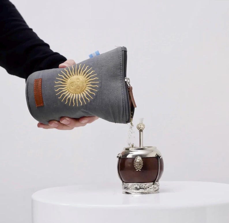 Op de afbeelding: Een grijze etui met een gouden zonnembleem giet inhoud in een bruine en zilveren yerba mate kalebas. De kalebas staat op een wit oppervlak. De etui heeft een bruin leren patch en een rits.