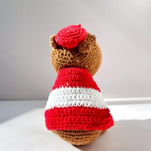 Op de afbeelding: Een handgemaakte bruine gehaakte beer met een rode baret en een rood-wit gestreept shirt. De beer is gehaakt met een getextureerde steek en de baret zit op zijn hoofd. De beer wordt van achteren bekeken.