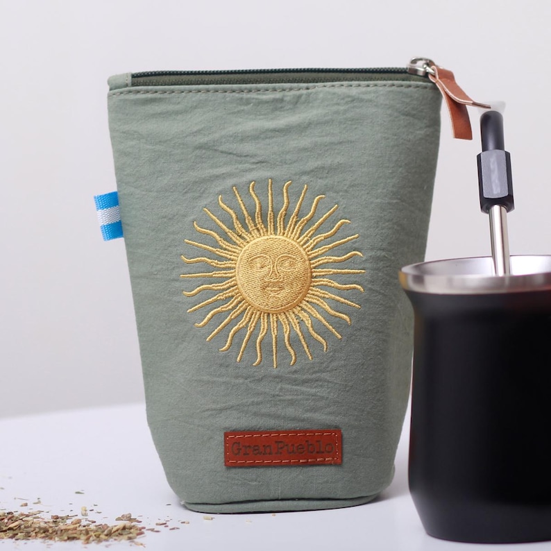 Op de afbeelding: Saliegroene stoffen etui met een gouden geborduurde zon. De etui heeft een ritssluiting en een klein leren treklipje. Een klein label met de tekst "Gran Pueblo" staat onderaan. Een zwarte beker en een rietje staan ernaast.