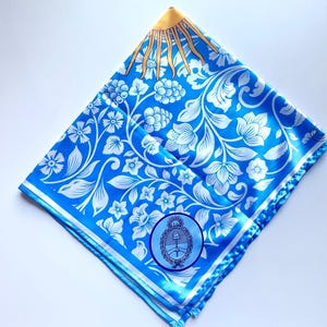 May include: A square, folded blue scarf with a white floral pattern and a gold sun design in the corner. The scarf also features a circular emblem with a coat of arms.