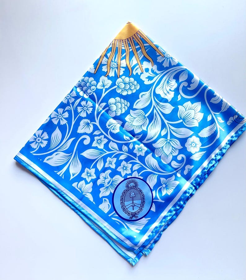 May include: A square, folded blue scarf with a white floral pattern and a gold sun design in the corner. The scarf also features a circular emblem with a coat of arms.