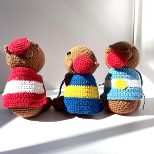 Op de afbeelding: Drie handgemaakte gehaakte poppen met bruine lichamen, rode neuzen en verschillende outfits. E&eacute;n pop draagt een rood-wit gestreept shirt, een andere een blauw-geel gestreept shirt en de derde een blauw-wit gestreept shirt.