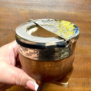 Może przedstawiać: Brązowy kubek do yerba mate z obramowaniem i pokrywką w kolorze srebrnym. Pokrywka ma ż&oacute;łto-niebieski wz&oacute;r. Kubek ma ozdobny srebrny pasek i czarny zamek błyskawiczny. Kubek spoczywa na trzech małych n&oacute;żkach.