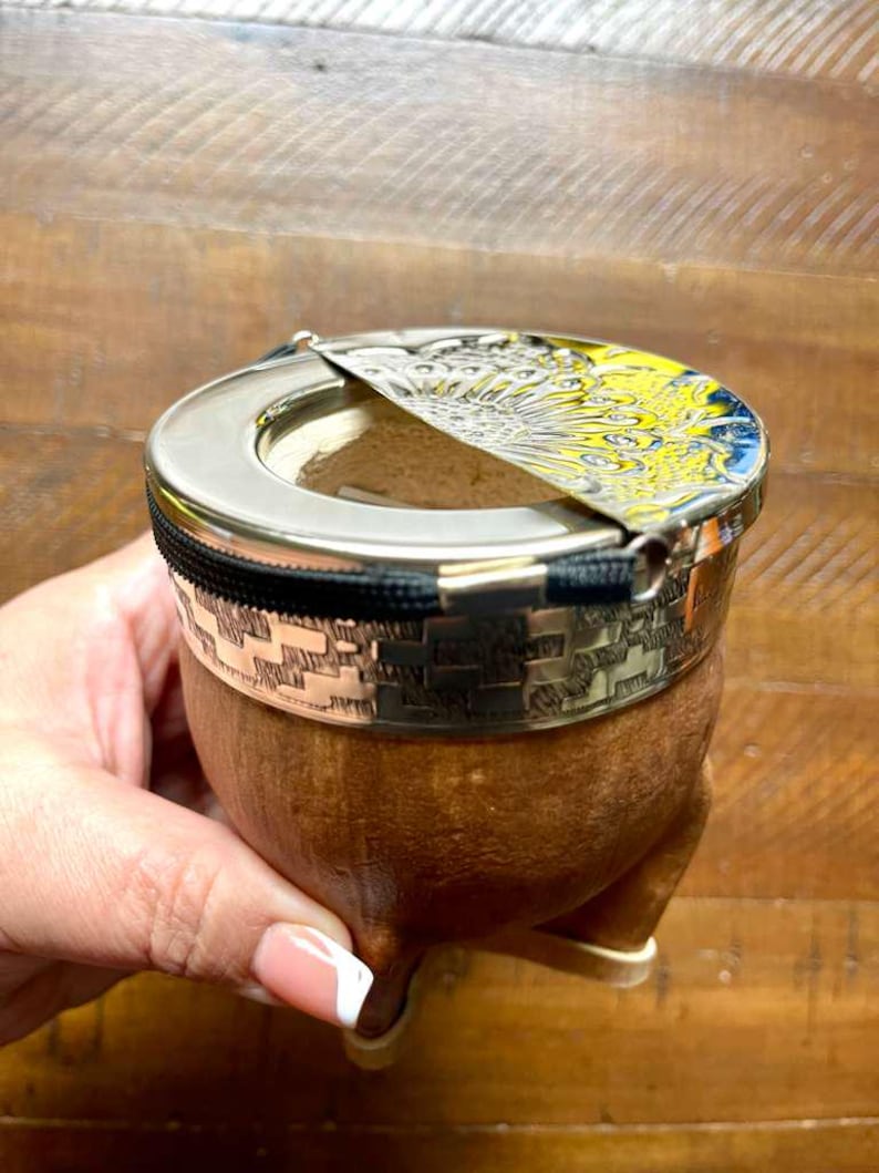 Może przedstawiać: Brązowy kubek do yerba mate z obramowaniem i pokrywką w kolorze srebrnym. Pokrywka ma ż&oacute;łto-niebieski wz&oacute;r. Kubek ma ozdobny srebrny pasek i czarny zamek błyskawiczny. Kubek spoczywa na trzech małych n&oacute;żkach.