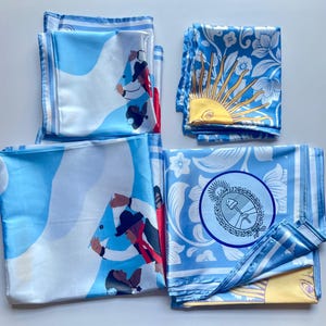 May include: Several folded silk scarves in shades of light blue and white are displayed. The scarves feature various designs, including illustrations of people, floral patterns, and a sun with a face. The edges of the scarves have a striped border.