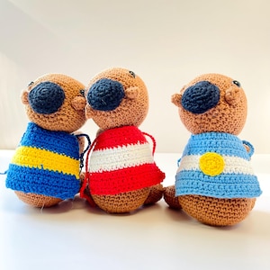 Op de afbeelding: Drie handgemaakte gehaakte otterpoppen. Elke otter heeft een bruin lichaam, een donkerblauwe neus en een bijpassende donkerblauwe hoed. De otters dragen truien in verschillende kleuren: blauw en gele strepen, rood en witte strepen en blauw en wit met een gele cirkel.