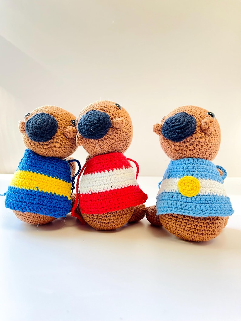 Op de afbeelding: Drie handgemaakte gehaakte otterpoppen. Elke otter heeft een bruin lichaam, een donkerblauwe neus en een bijpassende donkerblauwe hoed. De otters dragen truien in verschillende kleuren: blauw en gele strepen, rood en witte strepen en blauw en wit met een gele cirkel.