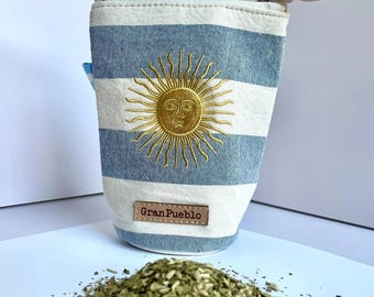 Puede incluir: Una bolsa cilíndrica con rayas azules y blancas, con un diseño de sol bordado en oro. La bolsa tiene un cierre de cremallera y una pequeña etiqueta de cuero. Un montón de hojas verdes secas se encuentra frente a la bolsa. La bolsa tiene el texto "Gran Pueblo".