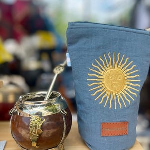 Peut inclure: Une calebasse à maté avec une base en métal et une paille bombilla en argent. À côté, une pochette bleue avec un motif de soleil brodé en or. La pochette porte une étiquette en cuir marron avec l'inscription "Para Pueblo".