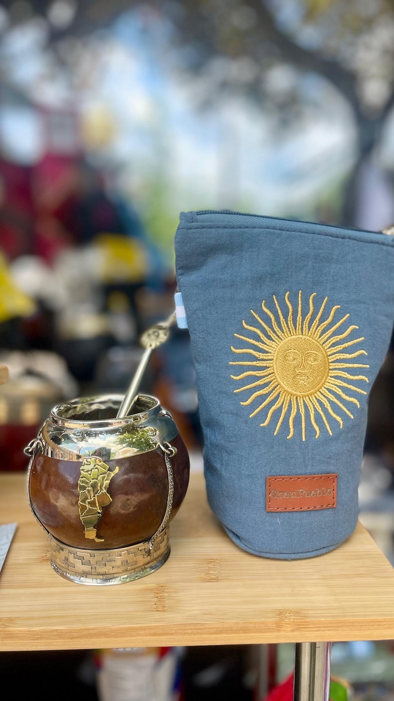Op de afbeelding: Een yerba mate kalebas met een metalen basis en een zilveren bombilla-rietje. Daarnaast een blauw zakje met een goud geborduurd zonontwerp. Het zakje heeft een bruin leren label met de tekst "Para Pueblo".