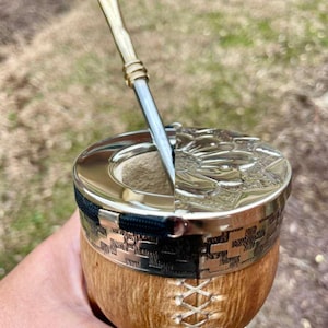 Może przedstawiać: Ręcznie wykonana tykwa do yerba mate ze srebrną obw&oacute;dką i pokrywką, z misternym wytłoczonym wzorem. Tykwa ma naturalne drewniane wykończenie i jest połączona z metalową słomką.