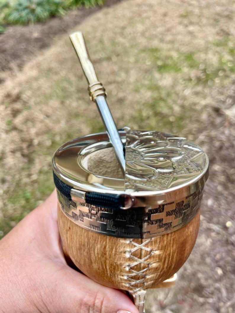 Może przedstawiać: Ręcznie wykonana tykwa do yerba mate ze srebrną obw&oacute;dką i pokrywką, z misternym wytłoczonym wzorem. Tykwa ma naturalne drewniane wykończenie i jest połączona z metalową słomką.