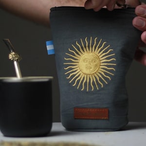 Op de afbeelding: Een donkergrijze etui met een goud geborduurd zonontwerp en een kleine Argentijnse vlag. Een bruin leren label is aan de onderkant genaaid. Een zwarte beker en metalen rietje zijn zichtbaar.