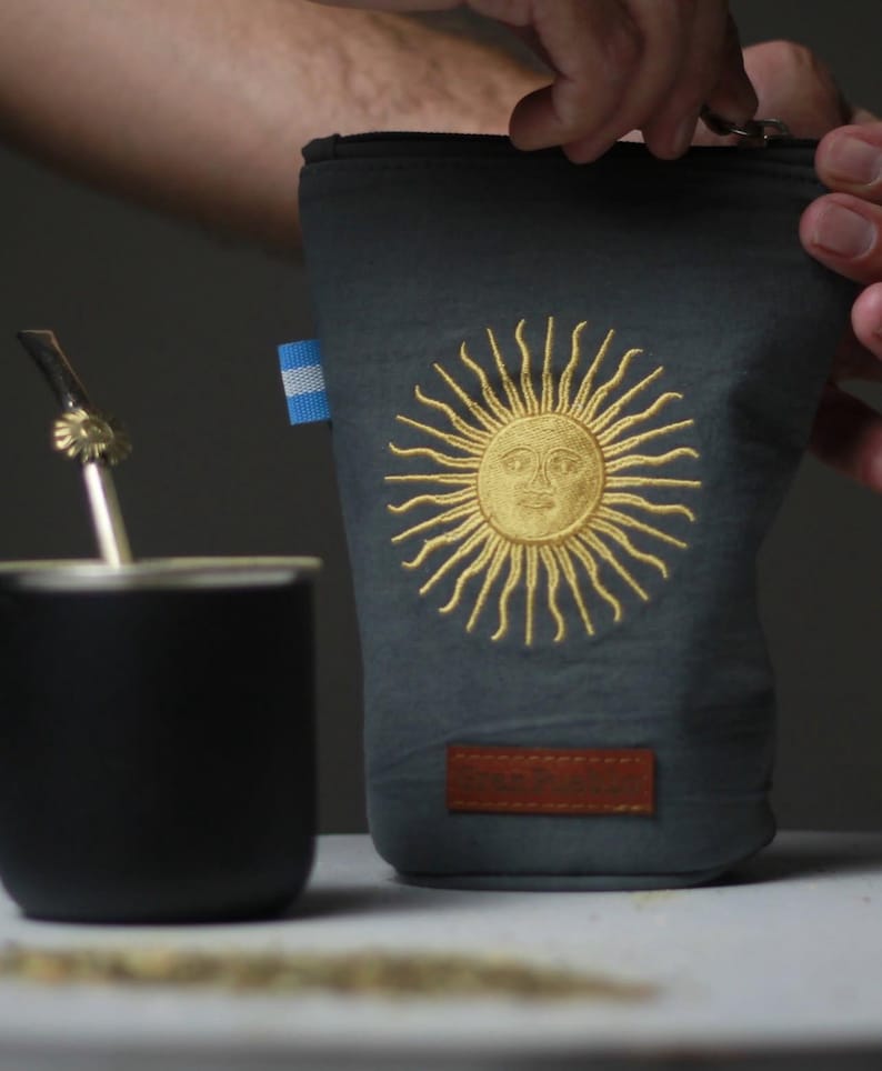 Op de afbeelding: Een donkergrijze etui met een goud geborduurd zonontwerp en een kleine Argentijnse vlag. Een bruin leren label is aan de onderkant genaaid. Een zwarte beker en metalen rietje zijn zichtbaar.