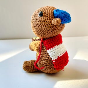 Op de afbeelding: Een handgemaakte bruine gehaakte pop met een blauwe hoed en een rood-witte gestreepte sjaal. De pop houdt een klein groen en bruin voorwerp vast. De pop zit. De pop heeft zwarte ogen en een klein oor.