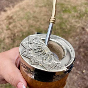 Może przedstawiać: Ręcznie wykonana tykwa do yerba mate ze zdobioną srebrną pokrywką z kwiatowym wzorem. Tykwa ma drewnianą podstawę i metalową słomkę. Tło jest rozmyte.
