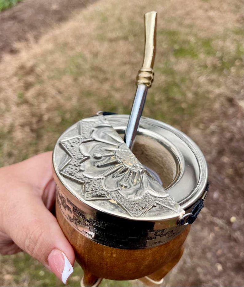 Może przedstawiać: Ręcznie wykonana tykwa do yerba mate ze zdobioną srebrną pokrywką z kwiatowym wzorem. Tykwa ma drewnianą podstawę i metalową słomkę. Tło jest rozmyte.