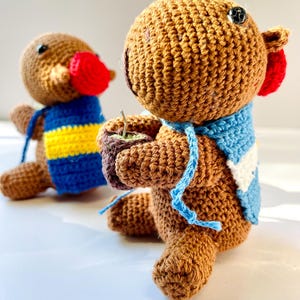Op de afbeelding: Twee handgemaakte gehaakte dierenfiguren. E&eacute;n figuur heeft een rode snavel en een blauw-geel gestreept vest. De andere figuur heeft een blauw-witte sjaal en houdt een klein kopje vast. Beide figuren zijn bruin met zwarte knoopogen.