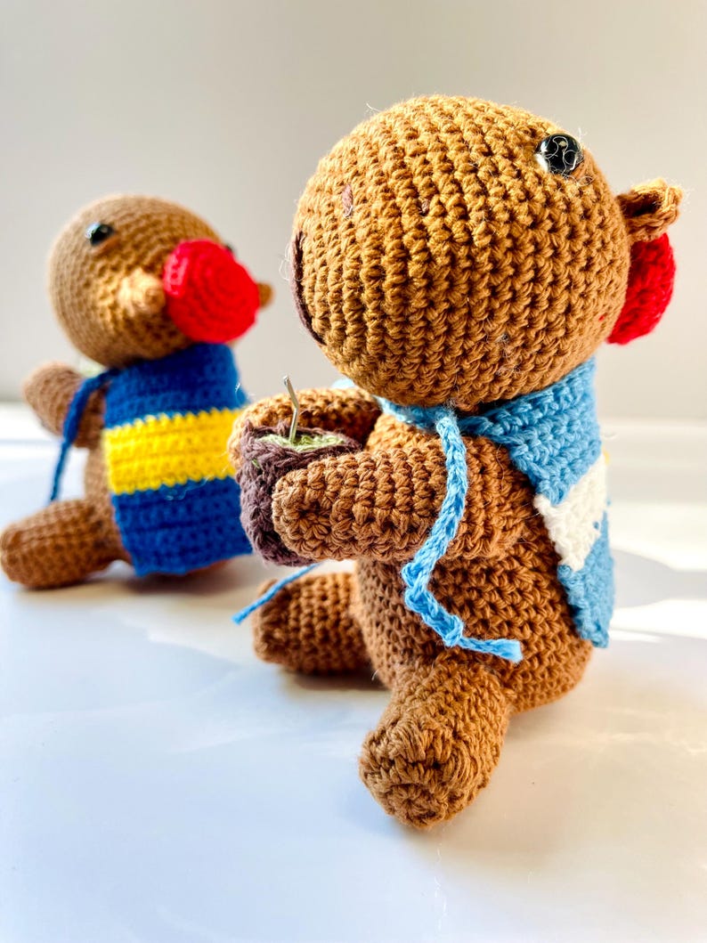 Op de afbeelding: Twee handgemaakte gehaakte dierenfiguren. E&eacute;n figuur heeft een rode snavel en een blauw-geel gestreept vest. De andere figuur heeft een blauw-witte sjaal en houdt een klein kopje vast. Beide figuren zijn bruin met zwarte knoopogen.