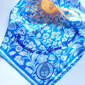 May include: A blue and white patterned scarf featuring a yellow sun with a face, floral designs, and the text "Coronados de Gloria Vivamos". The scarf also includes a circular emblem with a coat of arms.
