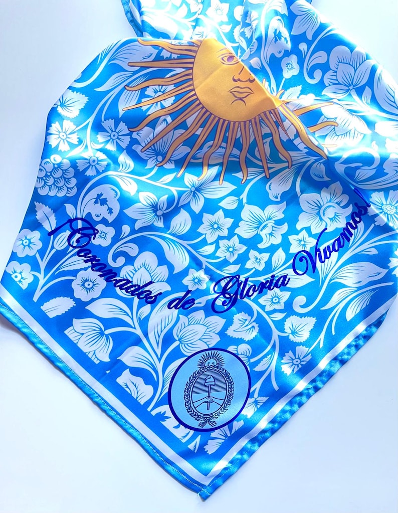 May include: A blue and white patterned scarf featuring a yellow sun with a face, floral designs, and the text "Coronados de Gloria Vivamos". The scarf also includes a circular emblem with a coat of arms.