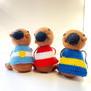 Op de afbeelding: Drie handgemaakte gehaakte otterpoppen. Elke otter heeft een bruin lichaam, een zwarte neus en draagt een trui. De truien zijn blauw en wit met een gele cirkel, rood-witte strepen en blauw-gele strepen.