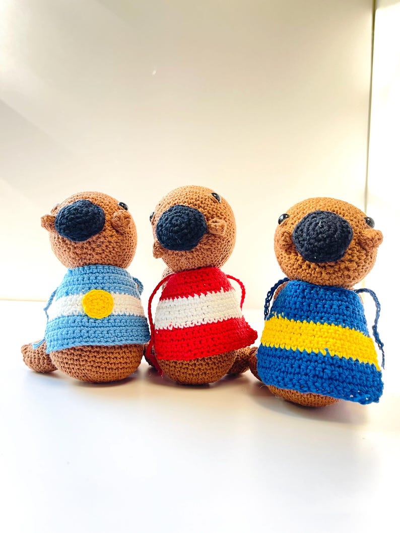 Op de afbeelding: Drie handgemaakte gehaakte otterpoppen. Elke otter heeft een bruin lichaam, een zwarte neus en draagt een trui. De truien zijn blauw en wit met een gele cirkel, rood-witte strepen en blauw-gele strepen.