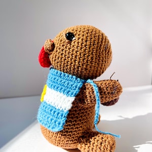 Op de afbeelding: Een handgemaakt bruin gehaakt dier met een rode neus en zwarte ogen. Het draagt een blauw-witte sjaal met een gele accent. Het speelgoed heeft een blauw touwtje.