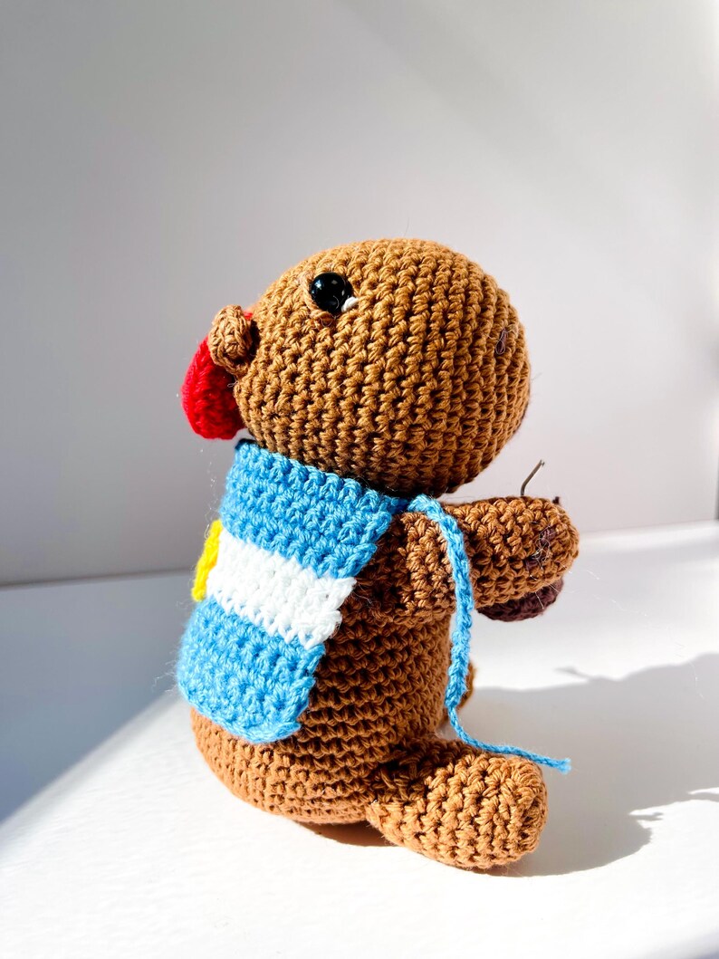 Op de afbeelding: Een handgemaakt bruin gehaakt dier met een rode neus en zwarte ogen. Het draagt een blauw-witte sjaal met een gele accent. Het speelgoed heeft een blauw touwtje.