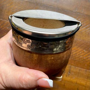 Może przedstawiać: Brązowy kubek do yerba mate w kształcie tykwy ze srebrnym metalowym obramowaniem i pokrywką. Obręcz ma wytłoczony geometryczny wz&oacute;r i jest zabezpieczona czarną opaską. Kubek trzymany jest w dłoni.