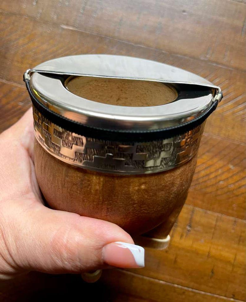 Może przedstawiać: Brązowy kubek do yerba mate w kształcie tykwy ze srebrnym metalowym obramowaniem i pokrywką. Obręcz ma wytłoczony geometryczny wz&oacute;r i jest zabezpieczona czarną opaską. Kubek trzymany jest w dłoni.