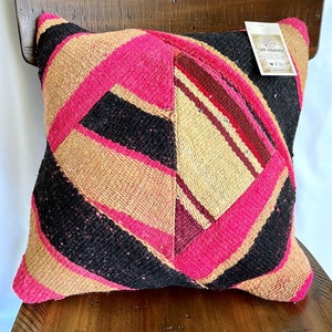 May include: A pink, black, and beige patterned throw pillow with a woven texture. The pillow has a tag attached that reads "VP Munay".