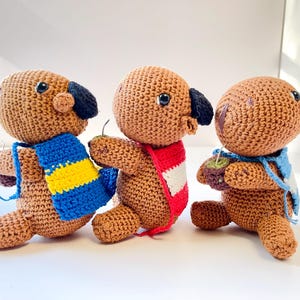 Op de afbeelding: Drie handgemaakte gehaakte hondpoppen in bruin met zwarte oren en blauwe ogen. Elke hond draagt een sjaal in een andere kleur: blauw en geel, rood en wit en blauw. De poppen staan alsof ze lopen.