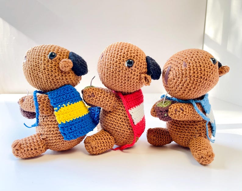 Op de afbeelding: Drie handgemaakte gehaakte hondpoppen in bruin met zwarte oren en blauwe ogen. Elke hond draagt een sjaal in een andere kleur: blauw en geel, rood en wit en blauw. De poppen staan alsof ze lopen.