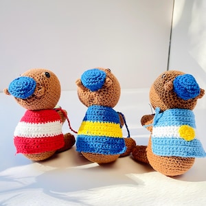 Op de afbeelding: Drie handgemaakte gehaakte vogelbekdierpoppen. Elke pop heeft een bruin lichaam, een blauwe snavel en een trui in een andere kleur: rood en wit gestreept, blauw met gele strepen en lichtblauw met een gele cirkel.