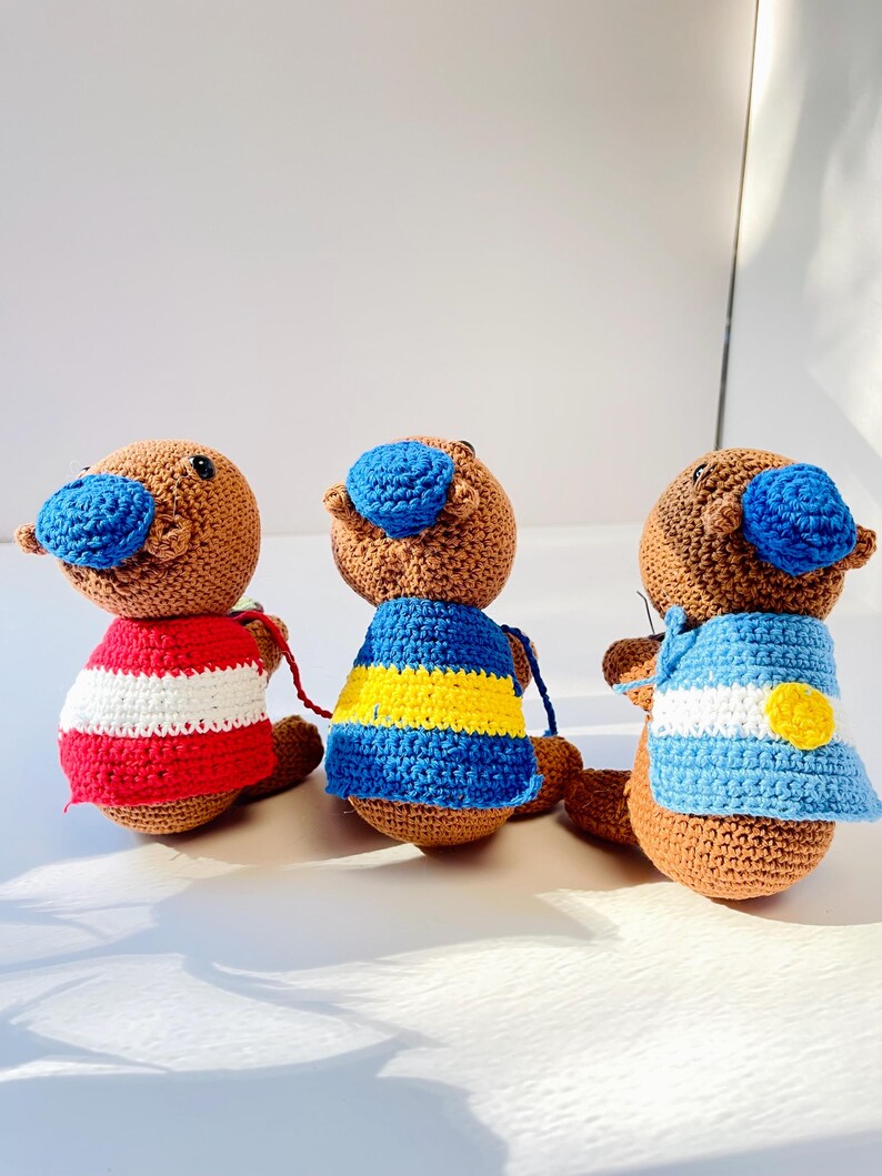 Op de afbeelding: Drie handgemaakte gehaakte vogelbekdierpoppen. Elke pop heeft een bruin lichaam, een blauwe snavel en een trui in een andere kleur: rood en wit gestreept, blauw met gele strepen en lichtblauw met een gele cirkel.