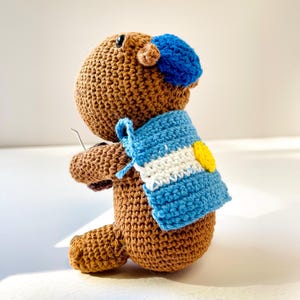 Op de afbeelding: Een handgemaakte gehaakte capibara-knuffel in bruintinten, met een blauwe baret en een blauw-witte sjaal met een gele accent. De knuffel is gemaakt met zichtbare steken, wat een getextureerd oppervlak laat zien.