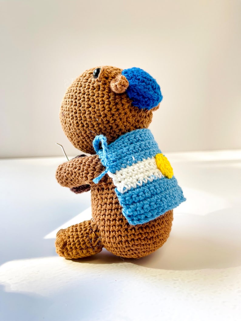 Op de afbeelding: Een handgemaakte gehaakte capibara-knuffel in bruintinten, met een blauwe baret en een blauw-witte sjaal met een gele accent. De knuffel is gemaakt met zichtbare steken, wat een getextureerd oppervlak laat zien.