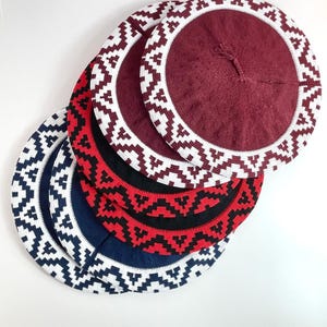 May include: A stack of four circular knit berets, each featuring a solid center and a wide border with a white and colored geometric zigzag pattern. The colors include deep burgundy, black, bright red, and navy blue, all with white accents.