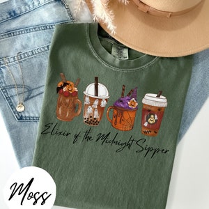 May include: A green t-shirt with a graphic of four Halloween-themed drinks. The text "Elixir of the Midnight Sipper" is printed below the drinks. The shirt is folded and displayed on a white surface.