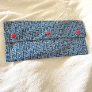 May include: A rectangular blue fabric pouch with a geometric white pattern and three red buttons. The pouch is closed with a flap and is laying on a white surface.