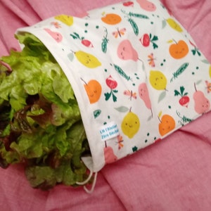 Peut inclure: Un sac à vrac en tissu blanc rempli de laitue fraîche. Le sac présente un motif répétitif de fruits et légumes colorés, notamment des poires, des citrons et des radis. Une petite étiquette blanche porte l'inscription LN l'Atelier Zéro Déchet.